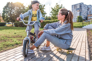 Happy laugh, Mom woman with little boy son of 4-5 years old, learn to ride bike, have fun enjoy relaxing, summer fall in city park area. Casual wear denim cap, sneaker pants. Skateboard are sitting.