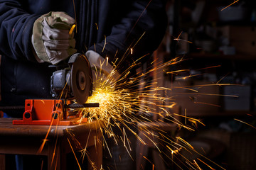 Hands of a craftsman with working gloves cutting an iron bar with the electric angle grinder which sprays many hot sparks in the dark workshop, dangerous work, copy space