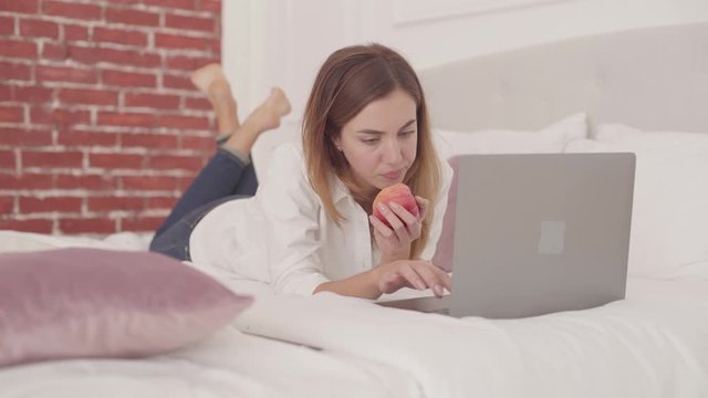 Young Caucasian woman laying in bed typing on laptop and eating red apple. Beautiful female student studying at home in casual atmosphere.