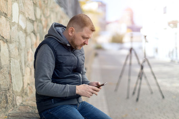 Male journalist is watching footage in his smartphone, sitting along the wall on the background of video tripods.