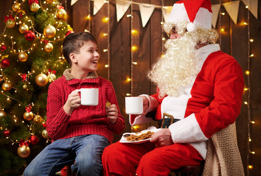 Santa Claus and child boy drinking tea, eating cookies, talking and sitting indoor near decorated xmas tree with lights - Merry Christmas and Happy Holidays!