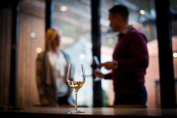 A glass of white wine on the table. The guy and the girl in the background talk