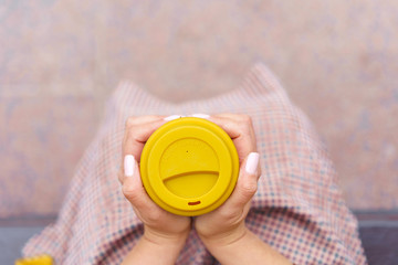 A woman is sitting on a park bench and holding a bamboo cup with hot coffee in her hands. On yellow silicone cap Caution contents hot. Fashionable autumn clothes in warm tones. View from above