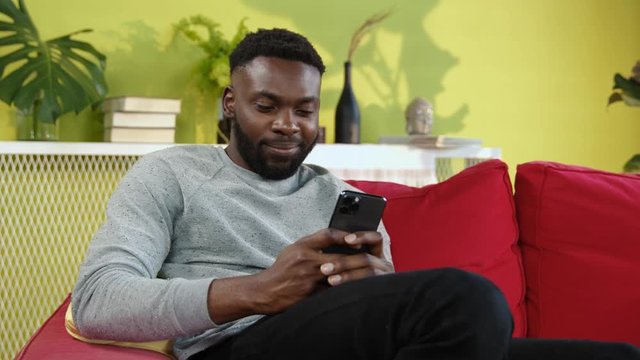 Cheerful young afro-american man browsing content in modern smartphone sitting on red sofa in stylish apartment living room.