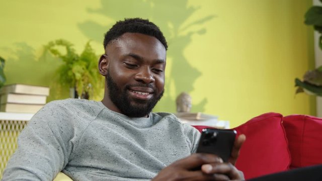 Cheerful afro-american guy laughing hen typing message to friend socializing on modern smartphone internet resting on red sofa at home.