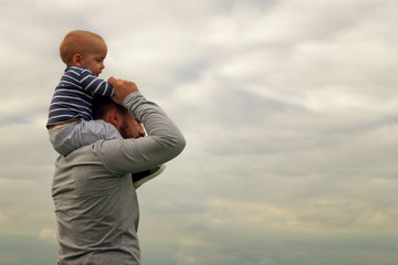 A child on his father’s neck. Walk near the water. Baby and dad against the sky.