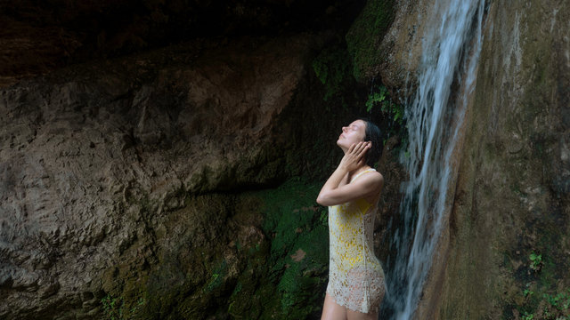 girl washes her face under the waterfall in the woods. Young woman in swimsuit in mountain river