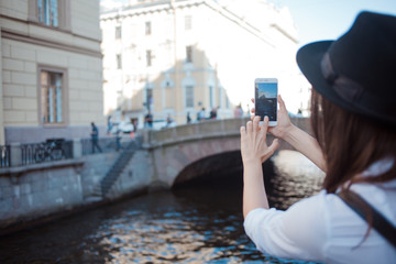 Tourist in the city takes a photo on smartphone. Young woman in black hat and white shirt.