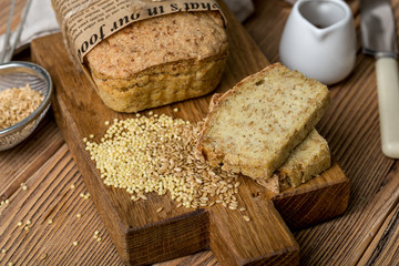 healthy content, abrick of freshly baked golden bread, couple of cut pieces, millet, flax, a scattering of grain, paper wrapper, wooden Board, textile, brown background, horizontal