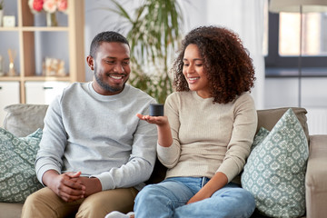 internet of things and technology concept - happy african american couple with smart speaker sitting on sofa at home