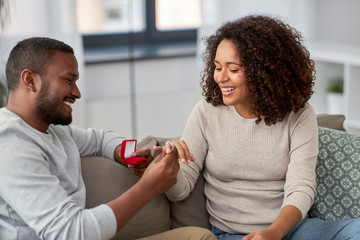 anniversary, proposal and couple concept - happy african american man giving diamond engagement ring in little red box to woman at home
