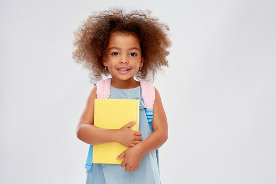 childhood, school and education concept - happy little african american girl with book and backpack over grey background