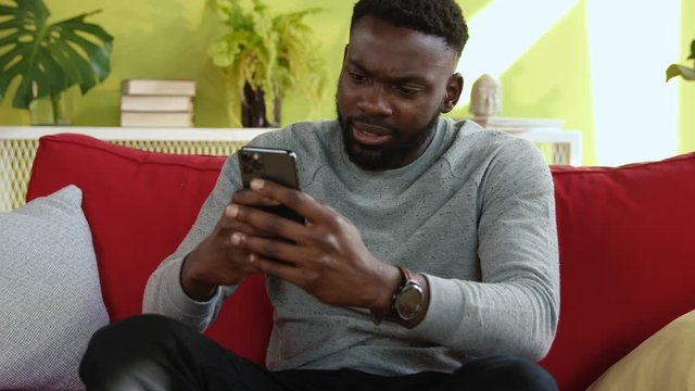 Afro-american man using modern smartphone resting on red sofa in the house. Excited handsome guy receiving great news cheering up and celebrating business success.