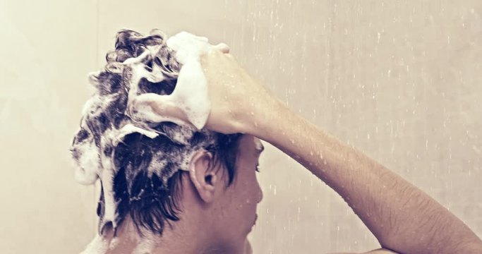 Rear view of an young man soaps hair with shampoo in the shower. Teenager takes shower in the bathroom. Man  washes under stream of warm water.   European human rinses hair with water. Slow motion.