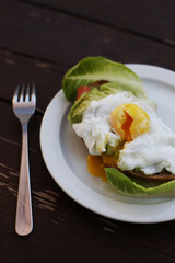 Close-up flat lay of delicious poached egg with liquid yolk on toast with fresh salad leaves and avocado at dark rustic brown wooden background/ table. Top view of healthy protein breakfast or lunch. 