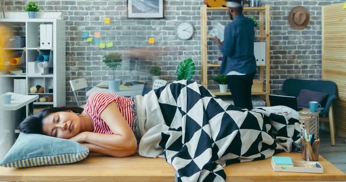 Zoom-in time lapse of female office worker sleeping on table in workplace relaxing while people colleagues are rushing in background. Lifestyle and youth concept.