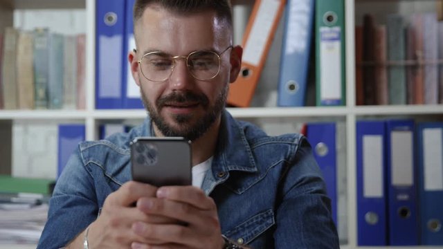 Portrait of nice-looking smiling young man using modern iphone browsing news messaging relaxing on break in the office. Business person with smartphone.