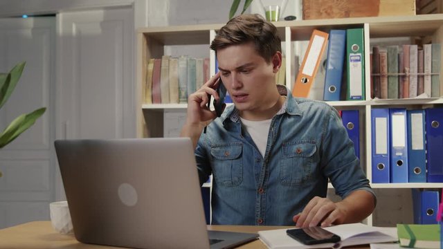 Busy young man office worker talking on two phones consulting customers managing multi-tasking deals working in company office.