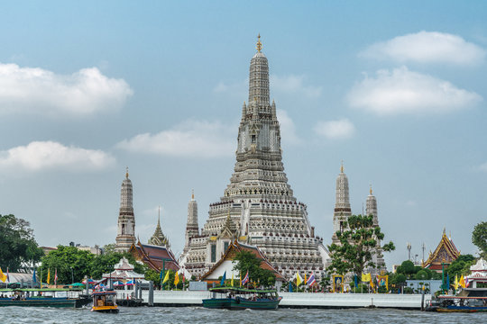 Bangkok city, Thailand - March 17, 2019: Side view on The spires or Prangs of Temple of Dawn against blue sky with some white clouds. Some green foliage, colorful flags and boats on Chao Phraya River 