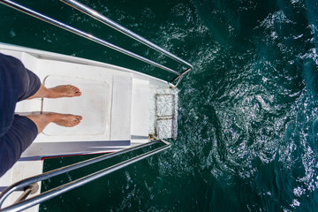 Fototapeta na wymiar The man's feet are on railing stairs for diving on a catamaran.Ladder of a sailing yacht to the sea with the raging sea water. Stairs with iron railings and white steps for swimming
