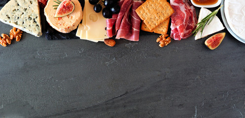 Assorted cheeses and deli meat appetizers. Top border, above view banner on a dark stone background...
