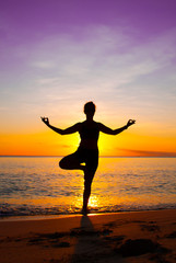 Fototapeta na wymiar A young lady practices yoga on the beach in front of the setting sun. The Caribbean sea is tranquil as it laps the sandy coastline of the idyllic island
