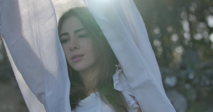Close-up portrair of a smiling Caucasian girl standing in the sunlight holding light white scarf in her raised hands. Beautiful woman spending summer evening outdoors.