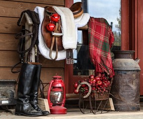 Portrait of a Black Great Dane resting on a farmhouse porch beside a saddle rack with English tack including saddle, bridle, riding boots decorated for Christmas- bells, lantern and plaid blanke - Powered by Adobe