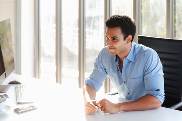 A short-haired Indian businessman wearing a blue shirt with a friendly, smiling face sitting at his office desk, with computers and mobile phones on the table, with a golden morning light.