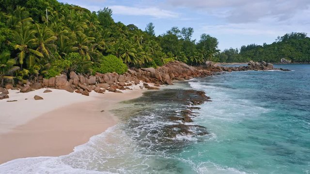 Aerial 4K low altitude fly over exotic tropical beach with rocks, palm trees and ocean waves at Mahe island, Seychelles