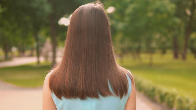 b roll rear female with long hair walks on pedestrian zone along alley with green trees brunette female strolls