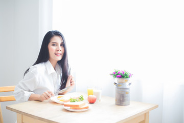 Obraz na płótnie Canvas A beautiful Asian woman, a tan, wearing a long-sleeved white shirt and shorts, and on her hand holding a fork and a knife, is eating breakfast in a bedroom at a hotel in Thailand