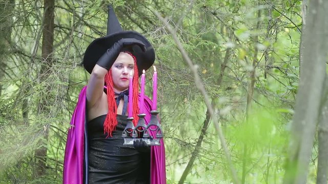 young witch with red hair and a black hat and a raspberry cloak conjures with candles in the forest.