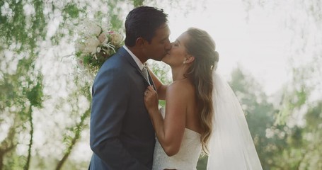 Happy young diverse couple kissing on their wedding day in nature with a backlit sun flare, amazing marriage moments in nature