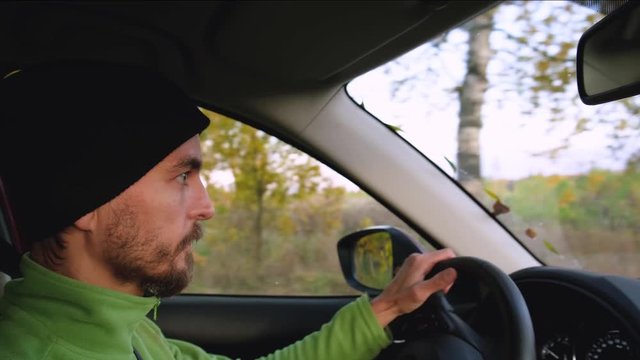 Young caucasian bearded man in knitted hat drives car on an autumn rural road with trees with   yellow foliage outside the window. Riadtrip or weekend journey concept. 