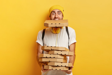 Portrait of overloaded pizza man holds many cardboard packages, one in mouth, wears casual white t...