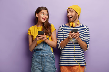 Joyful boyfriend and girlfriend laugh and look at each other, hold mobile phones, send text...