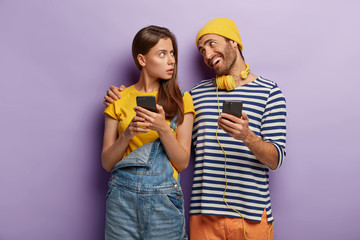Happy man embraces worried girlfriend, calms and talks pleasantly, use modern cell phones, check notifications, use bluetooth connection for sharing multimedia files, stand closely against purple wall