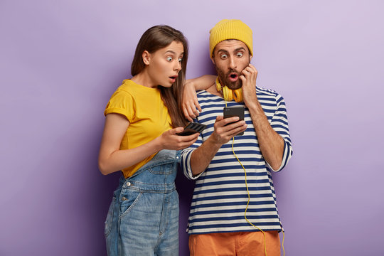 Scared teenage couple watch horror film in internet blog on modern cellular connected to wireless internet, stare at screen with omg expressions, being shocked to read horrible news on website