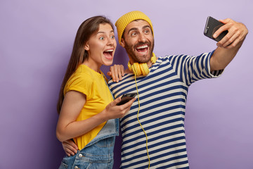 Two funny female and male friends take selfie on smartphone, have fun during recreation time, use modern mobile phone application, make common picture, dressed casually, isolated on purple wall