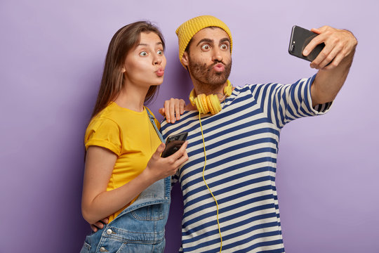 Funny young guy and woman make grimace, keep lips rounded, take picture on front camera of modern mobile phone, take selfie photos for social networks, spend free time together, stand indoor