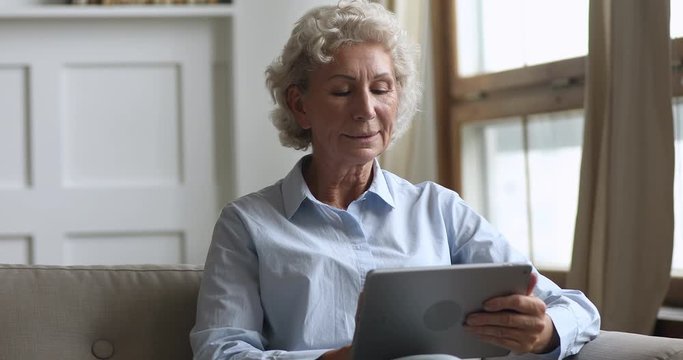 Happy old woman holding using digital tablet relax on sofa