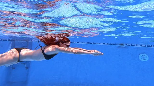 underwater view of redhead woman with black bikini and goggles swimming to breaststroke style in clear water of pool 