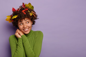 Cheerful dreamy Afro American girl tilts head, smiles pleasantly, looks aside, being in high spirit after autumn walk, makes portrait indoor, has yellow leaves, rowan berries in hair has friendly look