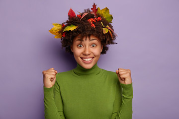 Image of lovely happy African American lady clenches fists from joy, smiles broadly, enjoys success, feels content and energized, celebrates achievement, enjoys autumn time, has leaves in hairstyle