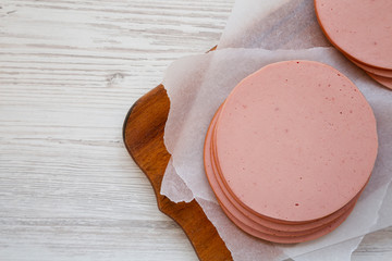 Sliced pork bolgna meat on a rustic wooden board on a white wooden surface, top view. Flat lay, overhead, from above. Copy space.