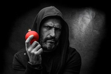 Black and white portrait of a bald bearded man in a hood with red christmas ball on a dirty gray background. Young Santa concept.