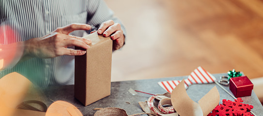 Woman sitting by the table and wrapping Christmas presents with brown paper