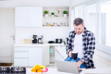 Man holding coffee and using a laptop in kitchen