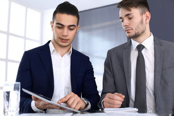 Two businessmen or partners working with tablet computer at the desk in modern office. Headshot of arab male entrepreneur or manager with colleague at workplace. Multi ethnic teamwork, partnership and